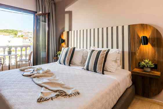 Ett hotellrum med en dubbelsäng, randiga prydnadskuddar och ett bord med stolar vid en öppen balkongdörr. (AI-genererad text)