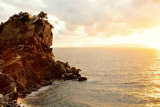 En klippa vid kusten med en byggnad och träd på toppen. Havet syns under en solnedgång. (AI-genererad text)