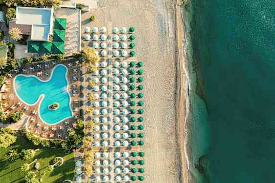 Flygfoto av en hotellpool med omgivande solstolar, bredvid en strand med parasoller och hav. (AI-genererad text)