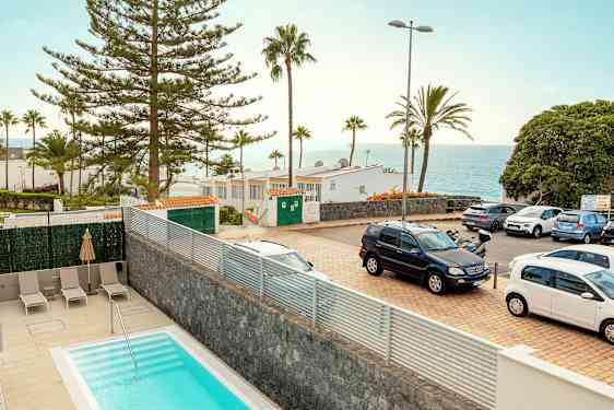 En bild av en utomhusmiljö med en pool i förgrunden, parkerade bilar, träd, byggnader och utsikt över havet i bakgrunden. (AI-genererad text)