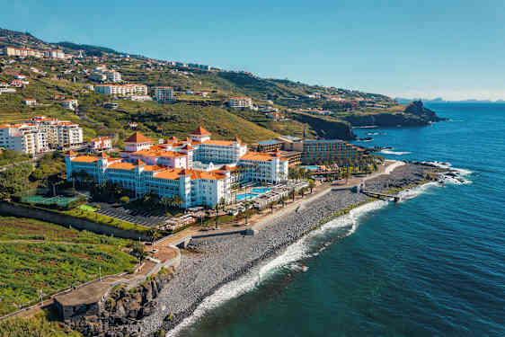 Ett hotell med röda tegeltak vid havet, omgiven av gröna kullar. (AI-genererad text)