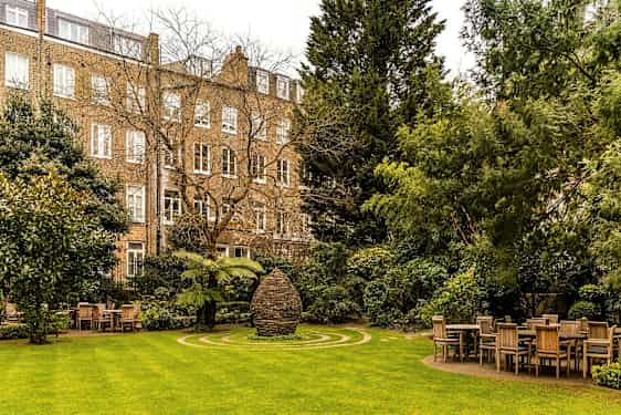 En grön trädgård med bord och stolar och ett stenmonument i mitten, omgiven av byggnader. (AI-genererad text)