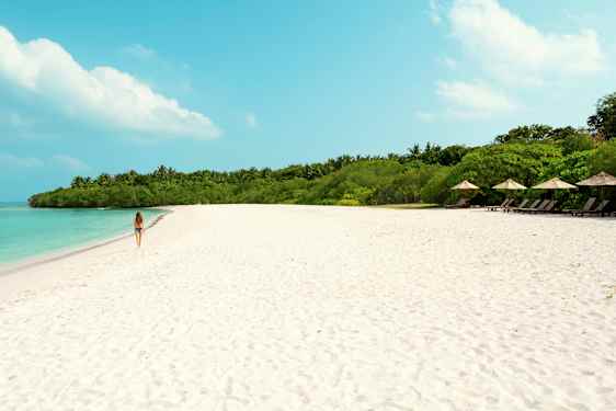 En kvinna går på en sandstrand med turkost vatten och grön vegetation i bakgrunden. (AI-genererad text)