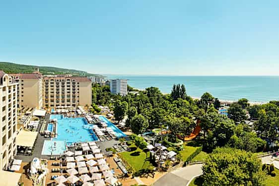 Flygbild av hotellkomplex med pooler, solstolar och grönska, beläget nära havet. (AI-genererad text)