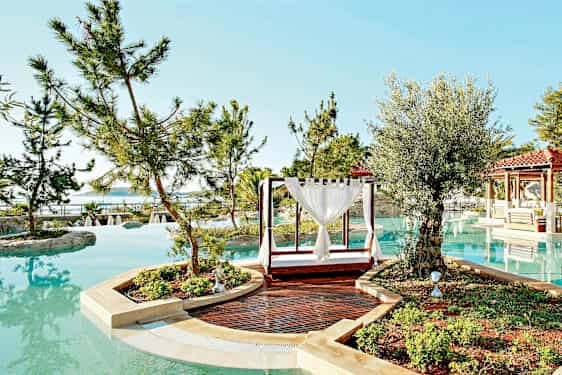 En pool omgiven av träd och växter med en utomhusdagbädd och en gångstig i förgrunden. (AI-genererad text)