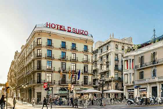 Ett gatuhörn med byggnader varav en är märkt som Hotel Suizo. Några människor går och sitter vid uteserveringar. (AI-genererad text)