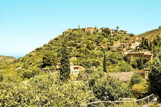 En scen med sluttande landskap fylld med grön vegetation, hus och låga byggnader utspridda bland träden. (AI-genererad text)