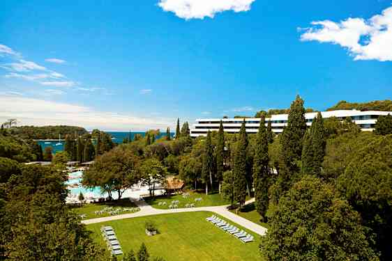 Hotellbyggnad omgiven av tallar och gröna fält med utsikt över havet. (AI-genererad text)