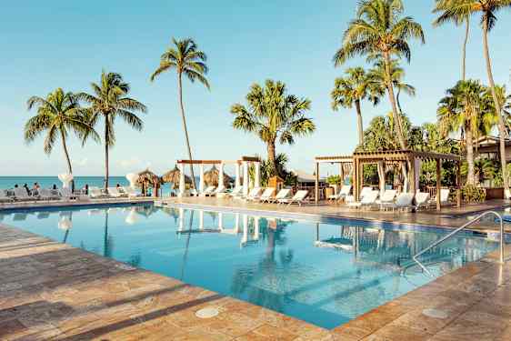 En pool omgiven av solstolar, palmer och en pergola, belägen nära havet med en klar blå himmel. (AI-genererad text)