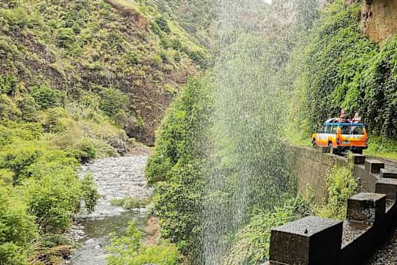 En gul-orange jeep på en väg vid ett vattenfall i en grön dal. (AI-genererad text)