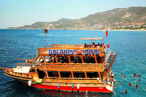 En träbåt med texten "Dolphin Dive" ligger vid klart blått vatten, där dykare syns i vattnet. I bakgrunden finns berg och en kustlinje. (AI-genererad text)