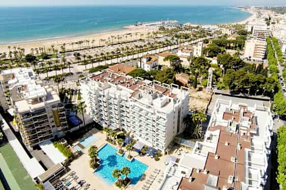 Luftbild av ett hotell med poolområde, omgärdat av byggnader och grönområden. I bakgrunden syns en sandstrand och hav. (AI-genererad text)