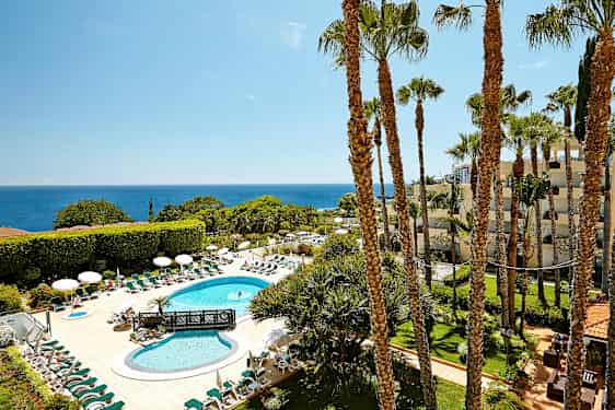 En hotellpool med solstolar och parasoller är omgiven av grönska med havet i bakgrunden. (AI-genererad text)