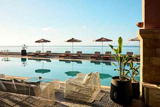 En swimmingpool med solstolar och parasoller vid sidan och utsikt över havet. Framför poolen står stolar och bord samt stora krukväxter. (AI-genererad text)