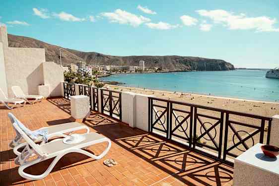 Terrass med solstolar och utsikt över en sandstrand med havet vid horisonten. (AI-genererad text)