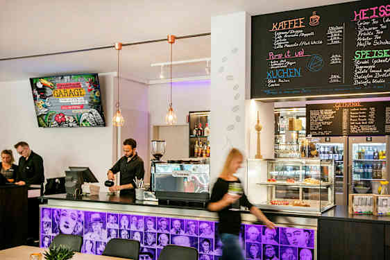 En caféinteriör med en disk som har kaffemaskin, bakverk i glasmonter och en meny på väggen ovanför. Personal står bakom disken. (AI-genererad text)