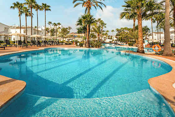 Stor utomhuspool med solstolar och parasoll omgiven av palmer och byggnader. (AI-genererad text)