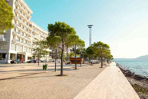 En strandpromenad med träd och vitkaklat golv längs vattnet, med en modern byggnad till vänster. (AI-genererad text)
