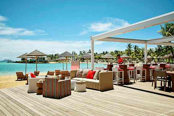 Uteservering vid stranden med bar och sittgrupper, samt utsikt över havet och omgivande palmer. (AI-genererad text)