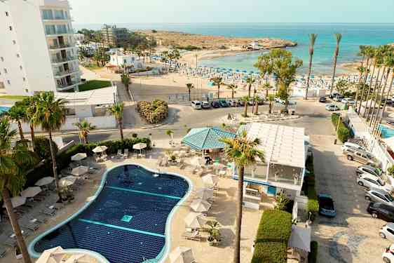 En översikt av en hotellpool med solstolar, omgiven av palmer, med havet och en solig strand i bakgrunden. (AI-genererad text)