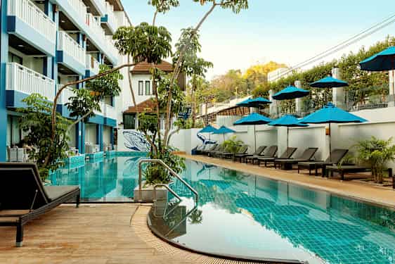 En utomhuspool med solstolar och parasoller, omgiven av vegetation och en hotellbyggnad i flera våningar. (AI-genererad text)