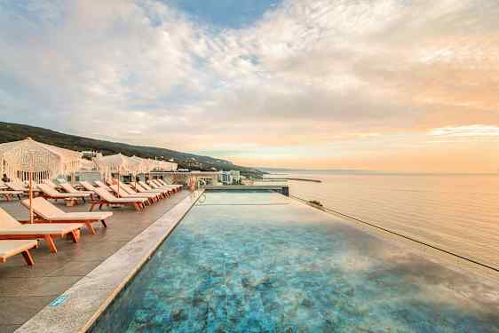 Utsikt över en infinitypool med solstolar och parasoller bredvid, mot havet i solnedgång. (AI-genererad text)