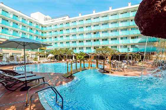 En utomhuspool vid en hotellbyggnad med solstolar, bord och parasoller. En liten bro över poolen och en del av en vattenfallstruktur syns. (AI-genererad text)