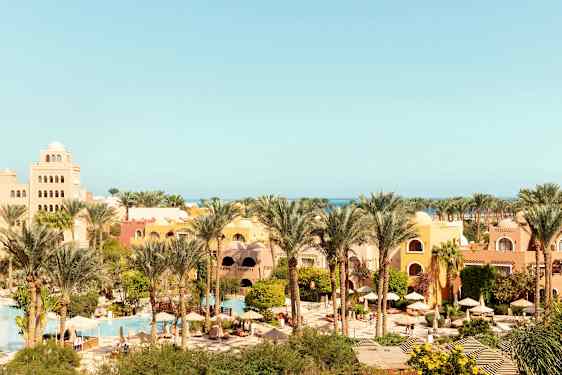 En semesterort med gula och orange byggnader, omgiven av palmer och grönska, med pooler och parasoller. (AI-genererad text)