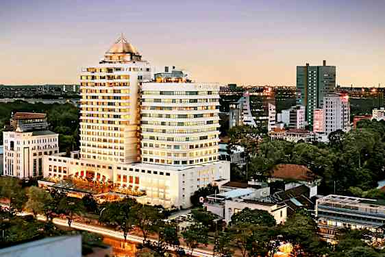 Vy över en stad under kvällstid med en stor byggnad i centrum, omgiven av mindre byggnader och träd. (AI-genererad text)