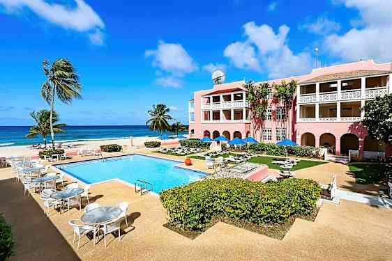 Bild av en solig resort med en utomhuspool, omgivande uteplatser och ett rosa hotell med balkonger, beläget vid havet. (AI-genererad text)