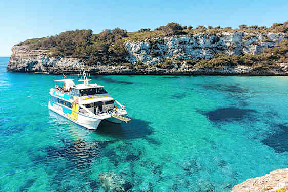 En vit båt med passagerare i en turkosblå vik, omgiven av klippiga kuster och grönska. (AI-genererad text)