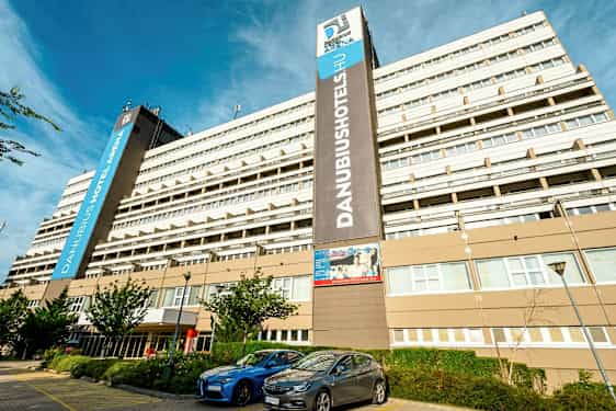 En stor hotellbyggnad med flera våningar med skylten ”Danubius Hotel Arena” synlig. Framför byggnaden finns parkerade bilar och några små träd. (AI-genererad text)