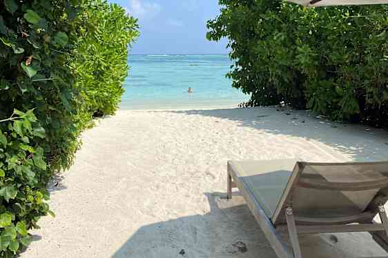 Solstol och parasoll på kritvit strand med grönska och turkosblått hav längre bort