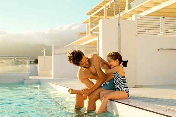 En liten flicka busar med en man vid poolkanten.