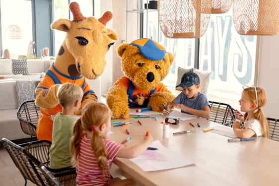 Lollo & Bernie går hand i hand med 3 barn, på en gångväg mellan lövträd. Fotografiet är taget bakifrån.