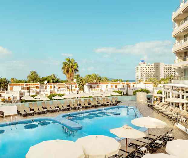 Utsikt över hotellpool med solstolar och parasoller, omgiven av byggnader och palmträd. (AI-genererad text)