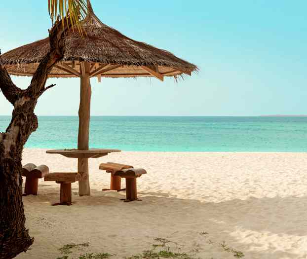 En parasoll med träbord och sittplatser på en sandstrand med havet i bakgrunden. (AI-genererad text)