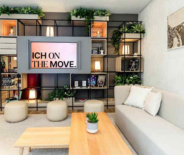 Ett modernt vardagsrum med hyllor fyllda av föremål och växtdekorationer, en soffa med kuddar, och ett soffbord med en krukväxt. (AI-genererad text)