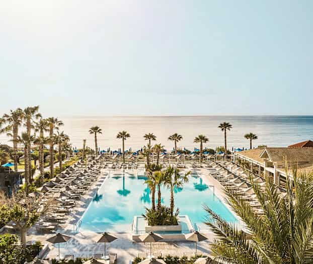 En stor utomhuspool omgiven av solstolar och parasoller med havet i bakgrunden. (AI-genererad text)