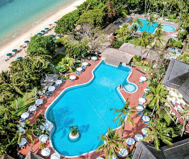 Flygbild av en poolanläggning omgivet av tropiska träd, med en strand och hav i bakgrunden. (AI-genererad text)