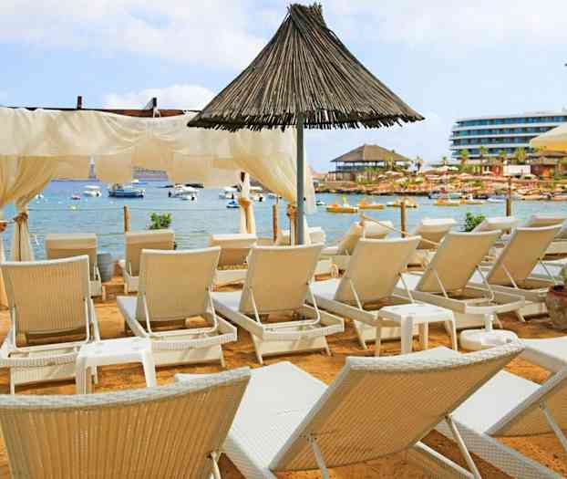 En strand med solstolar framför en lugn havsbukt med båtar. (AI-genererad text)