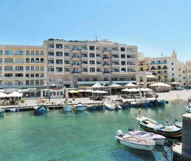 En bild av en hamn med båtar i klart vatten framför en rad av byggnader med uteserveringar och terrasser. (AI-genererad text)