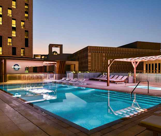 En upplyst utomhuspool på kvällen omgiven av solstolar och modern byggnadsarkitektur. (AI-genererad text)