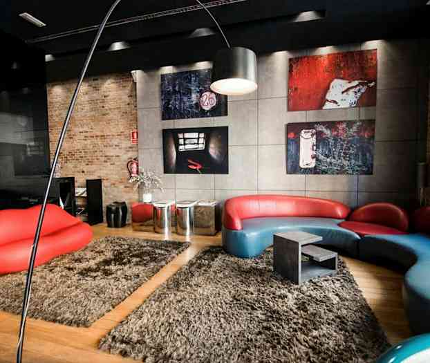 Ett modernt rum med en röd och blå soffa i unik design, en röd soffliknande sittmöbel formad som läppar, och konstverk på väggarna. Golvet är täckt av beige mattor. (AI-genererad text)