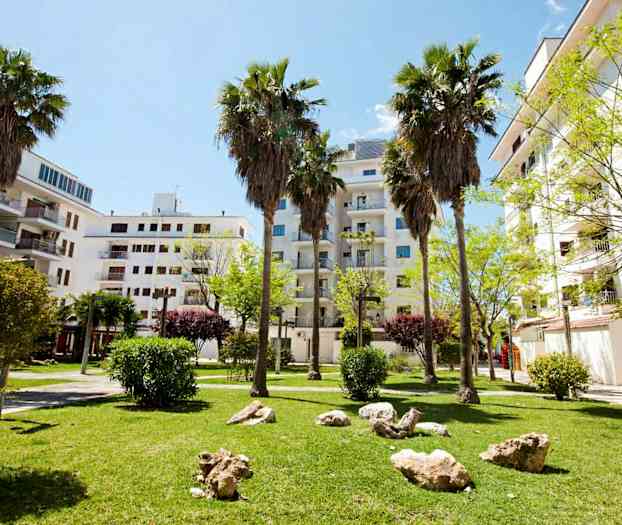 En grön park med palmer, gräsmatta och stenar, omgiven av vita byggnader. (AI-genererad text)