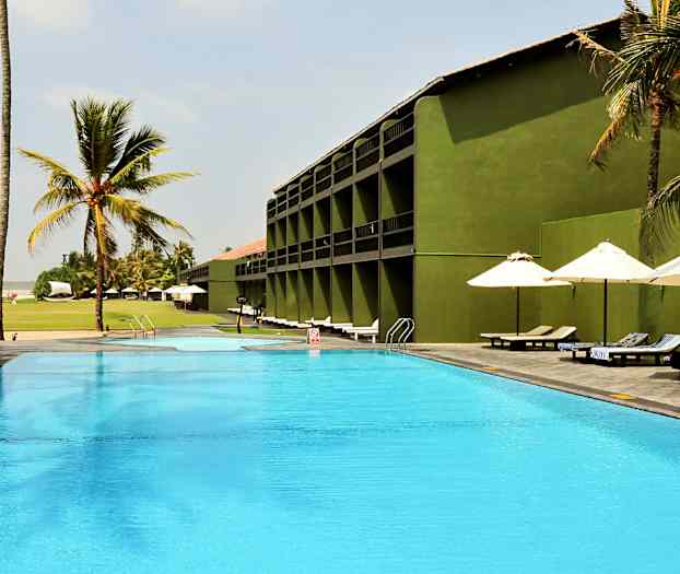 En utomhuspool vid ett hotell med gröna byggnader, omgiven av palmer och solstolar under parasoller. (AI-genererad text)