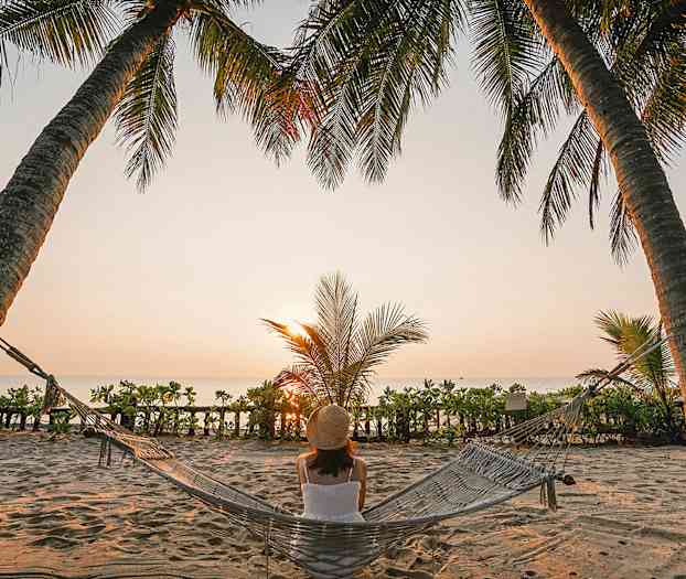 En person sitter i en hängmatta mellan två palmträd på en strand, medan solen går ner vid horisonten. (AI-genererad text)