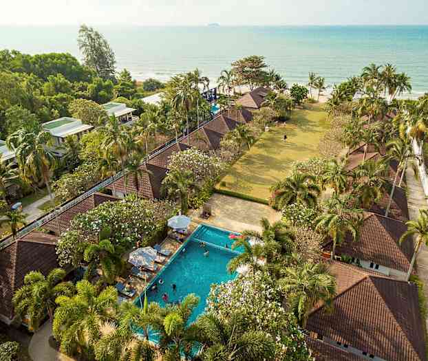 Flygbild över ett semesteränlägg med bungalower, pool, palmer och havsutsikt. (AI-genererad text)