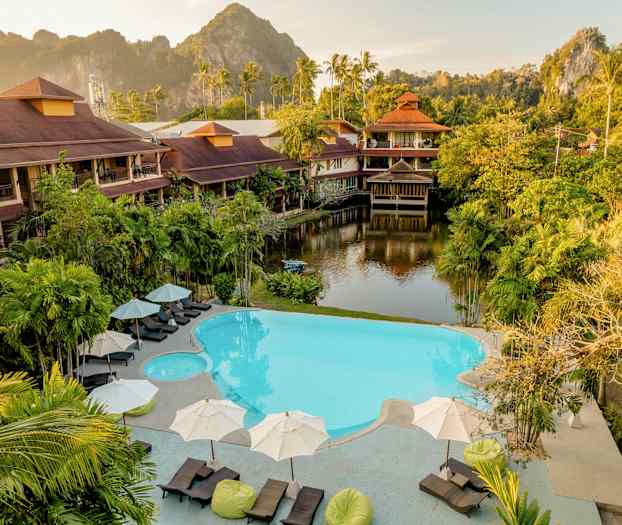 Ett hotellområde med poolområde omgivet av tropiska växter och byggnader i traditionell stil. I bakgrunden syns en grönskande bergssiluett. (AI-genererad text)