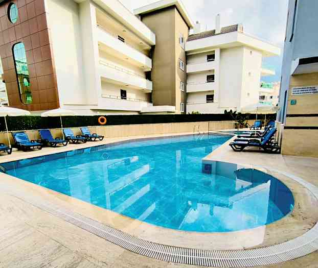 Utomhuspool med blått vatten omgiven av solstolar och byggnader. (AI-genererad text)